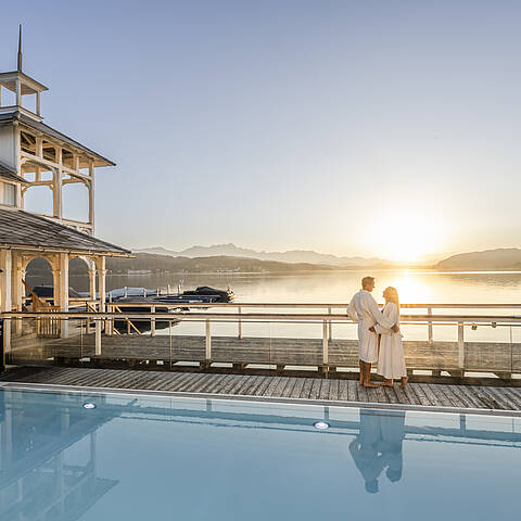 Paar im weißen Bademantel am Holzsteg vor dem beheizten Outdoorpool des Badehauses Werzers, mit Blick auf den Wörthersee bei Sonnenaufgang.